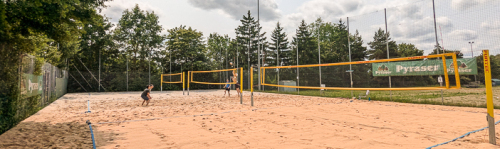4 Personen spielen Beachvolleyball auf einem Sandplatz im Freien. Das Spielfeld ist von einem Netz mit gelben Pfosten begrenzt, im Hintergrund stehen Bäume und ein Zaun. Eine Person bereitet sich darauf vor, den Ball anzunehmen. Der Himmel ist leicht bewölkt. © Post SV Nürnberg
