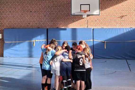 Eine Gruppe von Kindern steht eng im Kreis in einer Sporthalle, die Arme umeinander gelegt, unter einem Basketballkorb vor einer Backsteinwand.