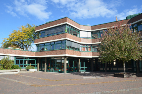 Mehrstöckiges Schulgebäude aus Backstein mit großen Fensterfronten und Eingangsbereich, davor ein gepflasterter Schulhof mit Bäumen unter blauem Himmel. © Jochen Schmid