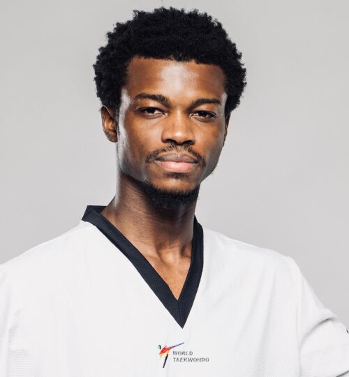 Porträt eines Mannes mit kurzem, schwarzem, lockigem Haar und leichtem Bart. Er trägt einen weißen Taekwondo-Anzug mit schwarzem Kragen vor hellem, neutralem Hintergrund.