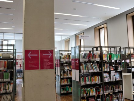 Das Bild zeigt den Bereich für Sprachen lernen auf der Ebene L1. Links und rechts im Bild sind Bücher·regale. In der Mitte vom Bild steht eine Säule. Auf der Säule sind 2 rote Schilder.