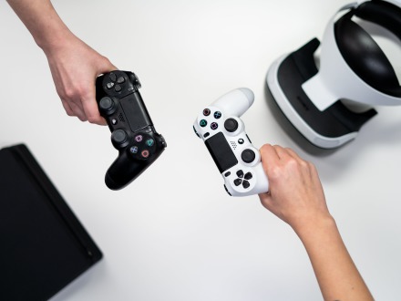 Das Bild zeigt 2 Hände. Eine Hand hält eine schwarze Spiele·konsole. Die andere Hand hält eine weiße Spiele·konsole. Auf der rechten Seite liegt noch eine weiße VR-Brille. Auf der linken Seite liegt ein schwarzer Kasten.