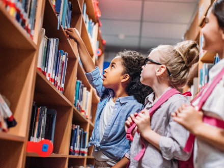 Das Bild zeigt: 3 Mädchen stehen vor einem Regal in einer Bibliothek. 1 Mädchen holt ein Buch aus dem Regal. Die anderen 2 Mädchen schauen zu.