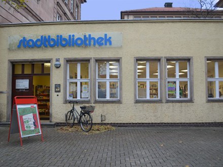 Das Bild zeigt den Eingang von der Stadt∙bibliothek Gostenhof. Vor der Tür ist eine Stufe. Rechts neben dem Eingang sind Fahrrad·ständer an der Wand. An dieser Wand sind mehrere Fenster. Vor dem Eingang steht ein Klapp·schild.