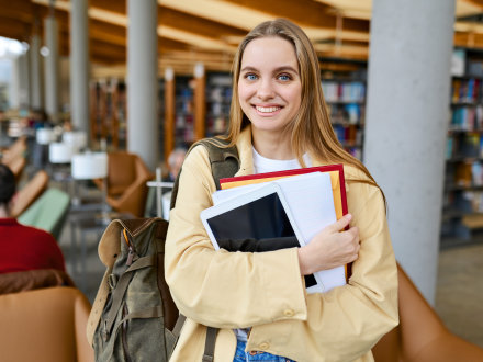 Das Bild zeigt eine lächelnde junge Frau in einer Bibliothek. Die junge Frau hält mehrere Hefte und ein Tablet im Arm.