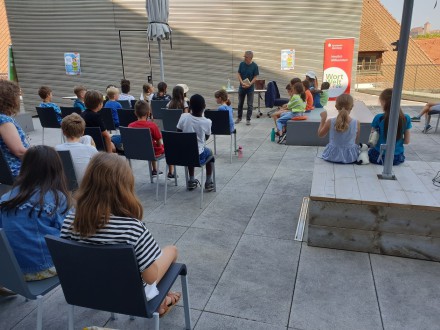 Das Bild zeigt eine Veranstaltung auf der Dach·terrasse. Viele Kinder sitzen auf Stühlen im Publikum. Vor den Kindern steht ein Mann. Der Mann redet zu den Kindern. Und der Mann hält ein Buch in der Hand.