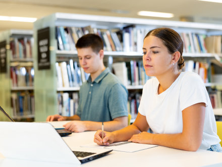 Das Bild zeigt 1 junge Frau und 1 jungen Mann an einem Arbeits·platz in einer Bibliothek.