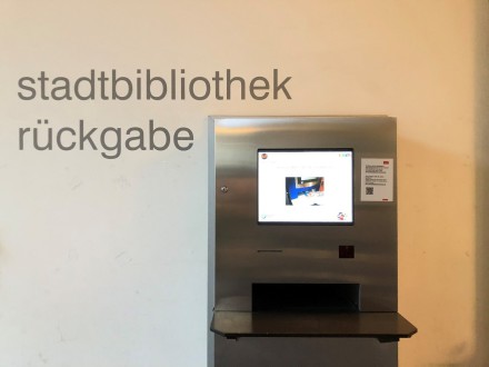 Dieses Bild zeigt einen Rückgabe∙automaten. Der Automat ist in einer Wand befestigt. Auf der Wand über dem Rückgabe∙automaten steht groß: stadtbibliothek rückgabe. Der Automat hat eine Ablage∙fläche und einen Bildschirm.
