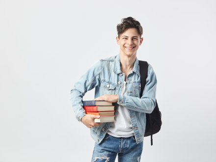 Das Bild zeigt einen jungen Mann in einer Jeans·jacke. Über der linken Schulter hängt ein schwarzer Rucksack. Der junge Mann hält 4 Bücher in den Händen.