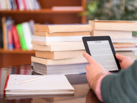 Das Bild zeigt einen E-Book-Reader. Eine Person hält den E-Book-Reader in den Händen. Daneben liegen auf einem Tisch 2 Bücher·stapel und 1 Block mit einem Stift.