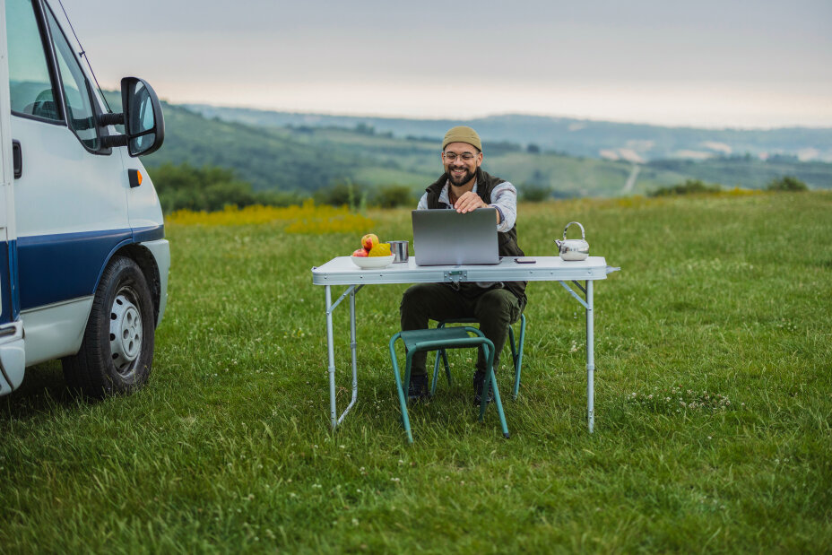 Happy male freelancer showing laptop screen while sitting at a table in front of his camper van, enjoying freedom and remote work opportunities in beautiful nature