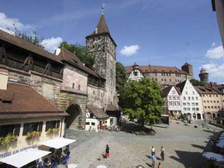 Foto von einem alten Markt·platz mit Kopfstein∙pflaster. Die Gebäude um den Platz sehen alt aus. Auf der linken Seite ist ein Turm zu sehen. Der Turm sieht ein wenig wie ein Kirch∙turm aus. Links neben dem Turm ist auch ein Bogen aus Stein als Durchgang.