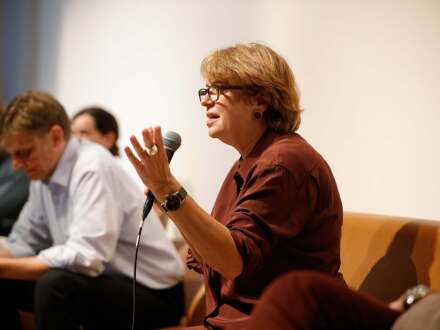Foto von einer Frau mit einem Mikrofon in der Hand. Die Frau spricht gerade. Neben der Frau sitzen noch andere Menschen und hören ihr zu.