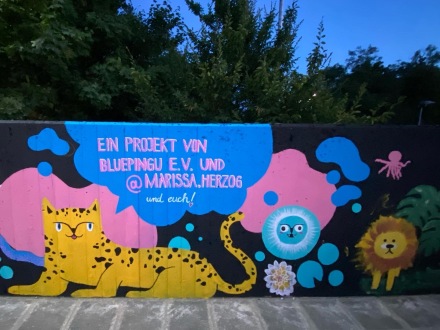 Foto von einer bunt bemalten Wand mit Tieren und bunten Formen. Zu sehen sind ein Leopard, ein Löwe und weitere bunte Formen. Über dem Leopard ist in eine blaue Sprech·blase gemalt. In der Sprech·blase steht in pinker und weißer Schrift: „Ein Projekt von Bluepingu e.V. und @marissa.herzog und euch!“ Hinter der Wand stehen einige Bäume. Unter dem Foto steht die Bild·unterschrift: SDGs go local – Bunte Wände für Nachhaltigkeit.
