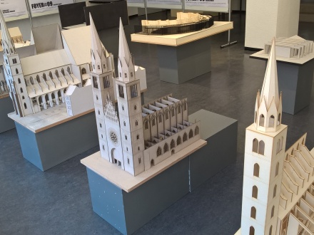 Foto von einer Ausstellung mit Modellen von alten Kirchen und Gebäude. Die Modelle sind aus hellem Holz. Die Modelle zeigen verschiedene Bauwerke. Die Modelle stehen auf grauen Sockeln. Im Hintergrund sieht man weitere Modelle.