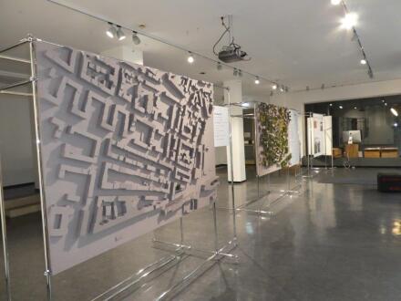 Foto von einem großen Raum. In dem Raum sind bei einer Ausstellung Fotos auf großen Leinwänden zu sehen. Das Foto im Vordergrund zeigt ein Stadt·modell. Der Raum ist hell beleuchtet.