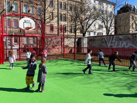 Foto von einem Ball·spielplatz. Auf dem Platz sind viele Kinder. Die Kinder spielen Fußball. Es gibt auch noch einen Basketball·korb.
