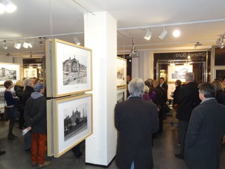 Foto von einer Gruppe von Menschen bei einer Ausstellung im Offenen Büro. Alte Fotos hängen an dünnen Drähten von der Decke. Die Besucher hören einer Person zu, die etwas über die Fotos erzählt. Der Raum ist hell beleuchtet.