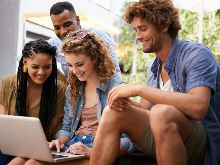 2 junge Frauen und 2 junge Männer sitzen zusammen. Eine Frau hat einen Laptop auf dem Schoß. Alle gucken lächelnd auf den Bildschirm von dem Laptop.