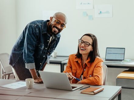 Foto von einer Frau und einem Mann in einem Büro. Die Frau sitzt an einem Schreib·tisch. Auf dem Schreib·tisch steht ein Laptop. Neben ihr steht der Mann und beugt sich leicht zu ihr herunter. Beide schauen zusammen auf den Bildschirm vom Laptop und lachen dabei.