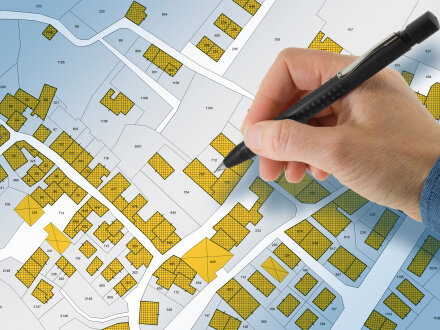 Foto von einem Bebauungs·plan. Der Bebauungs·plan zeigt einen Teil von einer Stadt. Auf dem Bebauungs·plan sind Gebäude in Form von gelben Kästchen eingezeichnet. Eine Person hält einen Stift in der Hand. Es wirkt, als möchte die Person etwas mit dem Stift auf dem Bebauungs·plan markieren.