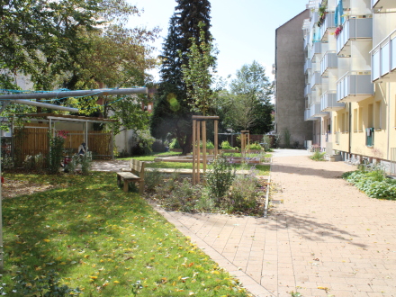 Das Foto zeigt den Hinter·hof von einem gelben Mehr·familien·haus. Am Haus ist der Hinter·hof mit hellen Steinen gepflastert. Links ist der Hinter·hof mit Bäumen und Sträuchern begrünt. Es gibt mehrere Rasen·flächen. Eine Holz·bank steht an so einer Rasen·fläche. Ein großer Wäsche·ständer steht auf der vordersten Rasen·fläche.