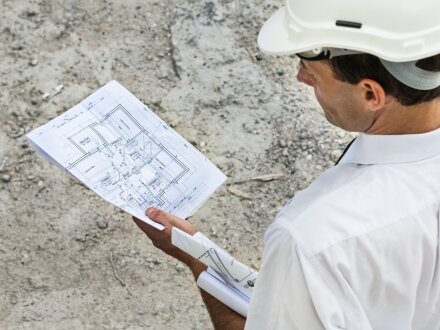 (Serge Nied Studio Chlorophylle / stock.adobe) Foto von einem Mann auf einer Bau·stelle. Der Mann trägt einen Schutz·helm auf dem Kopf. Der Mann prüft einen Bau·plan.