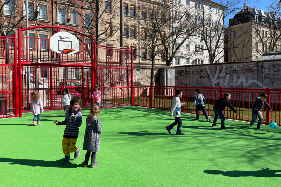 wird ergänztFoto von einem Ball·spielplatz. Auf dem Platz sind viele Kinder. Die Kinder spielen Fußball. Es gibt auch noch einen Basketball·korb.