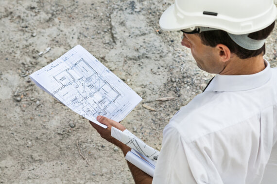 Foto von einem Mann auf einer Bau·stelle. Der Mann trägt einen Schutz·helm auf dem Kopf. Der Mann prüft einen Bau·plan.
