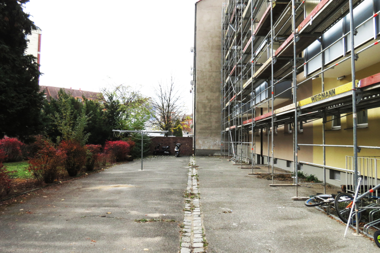 Foto von einem Hinter·hof von einem gelbem Mehr·familien·haus. An der Außen·wand vom Haus ist ein Gerüst aufgebaut. Der Hinter·hof hat eine graue Asphalt·fläche. Dort steht auch ein großer Wäsche·ständer. Links im Foto ist eine kleine Rasen·fläche mit ein paar Büschen und Bäumen.