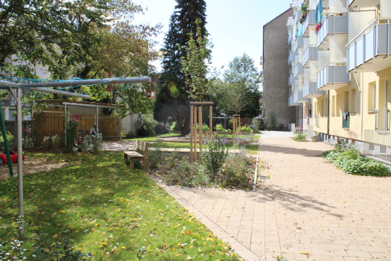 Das Foto zeigt den Hinter·hof von einem gelben Mehr·familien·haus. Am Haus ist der Hinter·hof mit hellen Steinen gepflastert. Links ist der Hinter·hof mit Bäumen und Sträuchern begrünt. Es gibt mehrere Rasen·flächen. Eine Holz·bank steht an so einer Rasen·fläche. Ein großer Wäsche·ständer steht auf der vordersten Rasen·fläche.