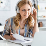 Eine Frau sitzt hinter einem aufgeklappten Laptop. Sie hat Kopfhörer auf und blättert in einem Spiralblock.