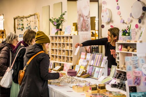 Besucher des Herzlmarkts sehen sich eine Auslage an. Eine Verkäuferin reicht ihnen eine handgefertigte Postkarte über den Verkaufstisch.