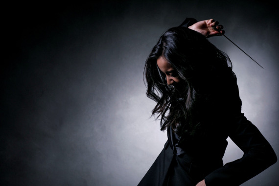 Porträt der Musikerin Erina Yashina. Sie trägt schwarze Kleidung. Ihr Gesicht wird von langen schwarzen Haaren verdeckt. Sie hält einen Dirigentenstab über dem Kopf. Der Hintergrund ist grau.