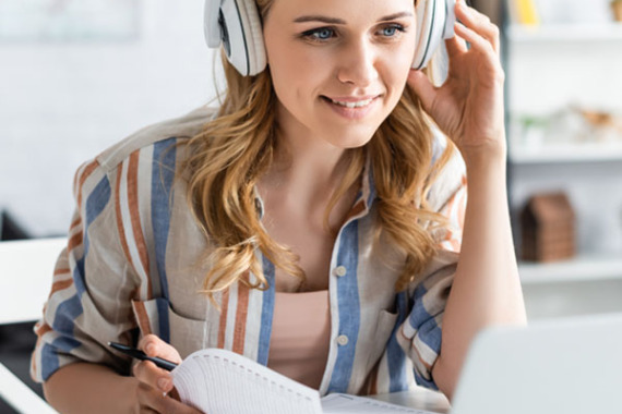 Foto von einer Frau. Die Frau sitzt hinter einem aufgeklappten Laptop. Sie hat Kopfhörer auf und blättert in einem Spiralblock.