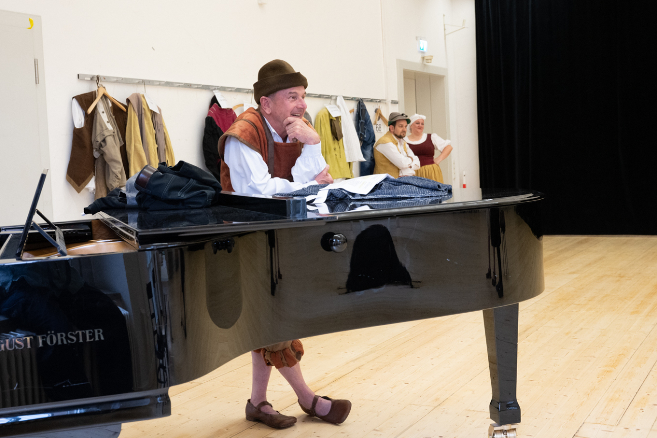 Foto: Mitglieder des Ensembles der Hans-Sachs Spielgruppe, schauen bei den Proben zu. Im Vordergrund steht ein schwarzer Flügel auf dem ein Schauspieler sich abstützt und lächelnd seinen Kollegen beim Proben zusieht. Im Hintergrund hängen Requisiten und Kostüme an der Wand., Bild © Christine Dierenbach/ Stadt Nürnberg / Stadt Nürnberg/Christine Dierenbach