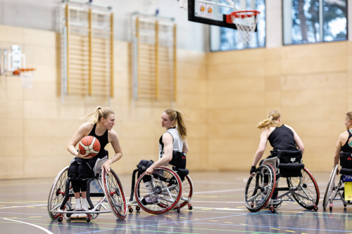 Foto: Rollstuhlbasketballerin Lilly Sellak inmitten eines Spiels. Sie hält in der rechten Hand den Basketball, mit der linken steuert sie ihren Rollstuhl. Drei weiter Mitspielerinnen sind zu sehen.