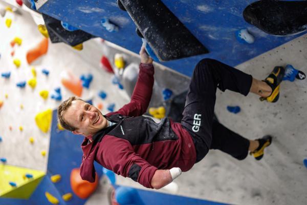 Foto: Parakletterer Kevin Bartke, hält sich mit dem linken Arm in der Kletterwand fest und lacht in die Kamera. Im Vordergrund sieht man den rechten Arm, bei dem der Unterarm fehlt .