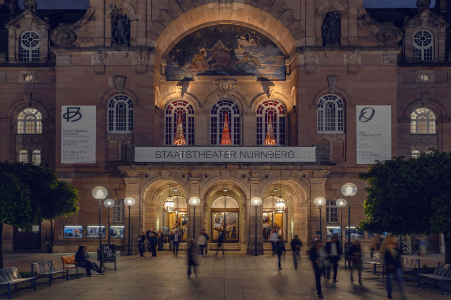 Foto: Fassade des Opernhauses in abendlicher Beleuchtung, davor unscharf Besucherinnen und Besucher., Bild © Staatstheater Nürnberg/Matthias Dengler