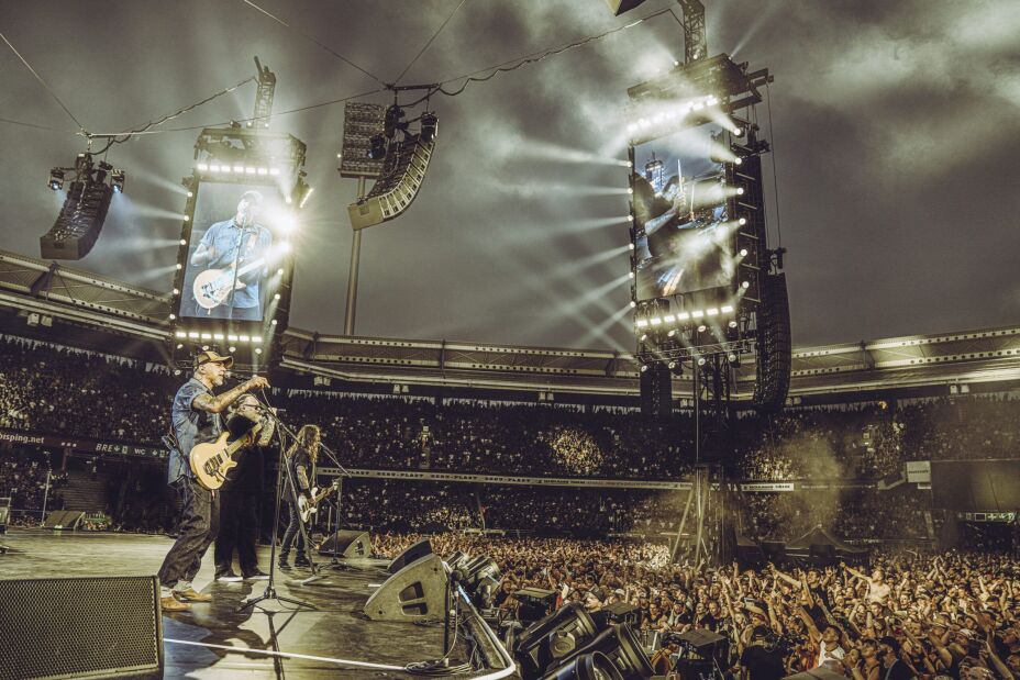 Foto: Voll besetztes Stadion mit abendlicher Beleuchtung, links Bühne mit drei Musikern mit Mikrofon, zwei davon mit Gitarren., Bild © Moritz Mumpi Künster