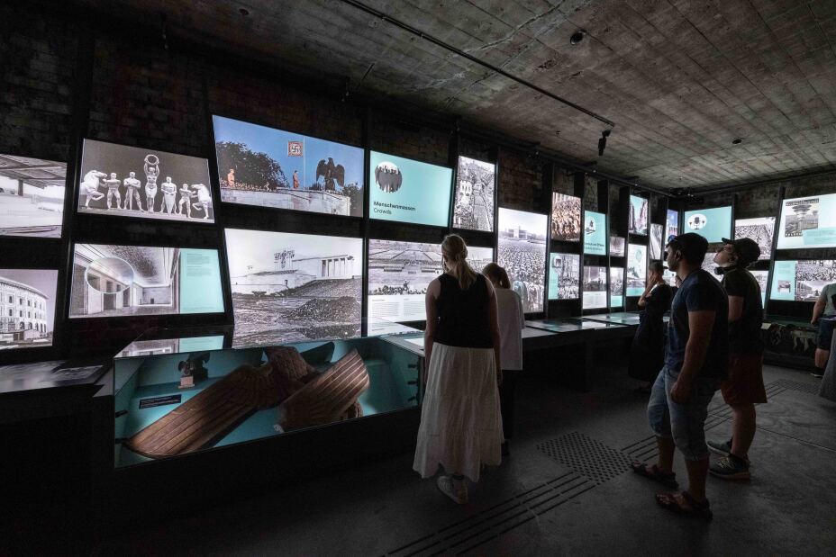 Foto: Blick in die Ausstellung des Dokumentationszentrums Reichsparteitagsgelände. Besucherinnen und Besucher vor beleuchteten Ausstellungstafeln und Vitrinen., Bild © Stadt Nürnberg/Christine Dierenbach