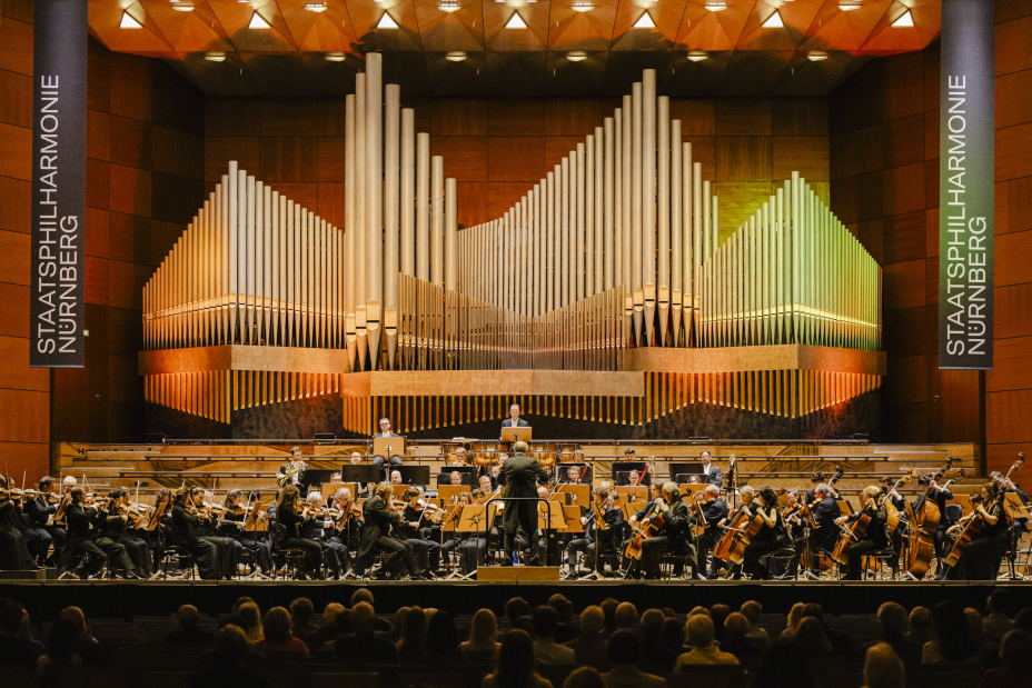 Foto: Bühne der Meistersingerhalle mit großem Orchester, im Hintergrund eine große Orgel, im Vordergrund Zuschauer von hinten., Bild © Staatsphilharmonie Nürnberg/Sebastian Lock