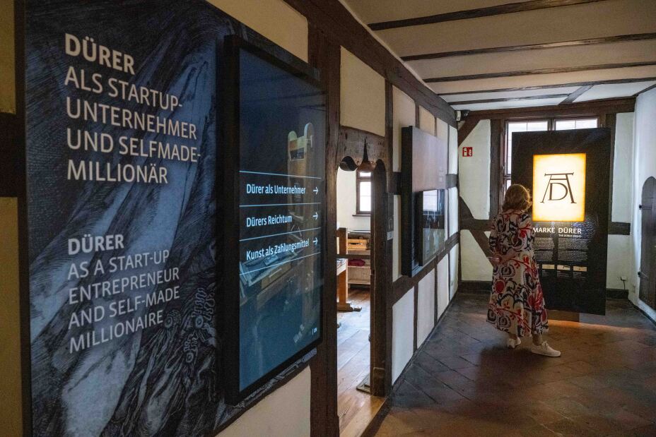 Bild © Stadt Nürnberg / Christine Dierenbach Foto: Blick in die Ausstellung des Albrecht-Dürer-Hauses. Auf der linken Seite Ausstellungstafeln, am Ende des Gangs eine beleuchtete Tafel mit dem Monogramm AD, davor eine Frau, die die Tafel betrachtet., Bild © Stadt Nürnberg / Christine Dierenbach