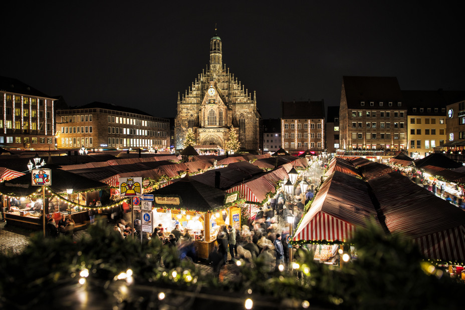 Foto: Weihnachtsmarkt schräg von oben mit abendlicher Beleuchtung, Menschen in den Gassen zwischen den Buden mit rot-weiß gestreiften Dächern, dahinter beleuchtete Kirchenfassade., Bild © floriantrykowski.com