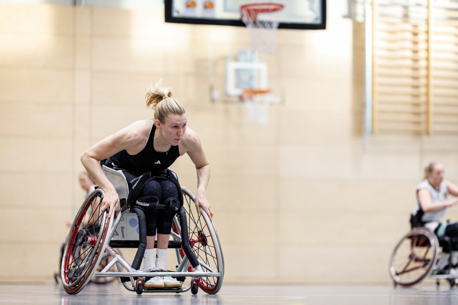 Foto: Rollstuhlbasketballerin  Lilly Sellak beim Basketballspiel in einer Sporthalle. Sie schiebt mit ihren muskulösen Armen kraftvoll ihren Stuhl an.