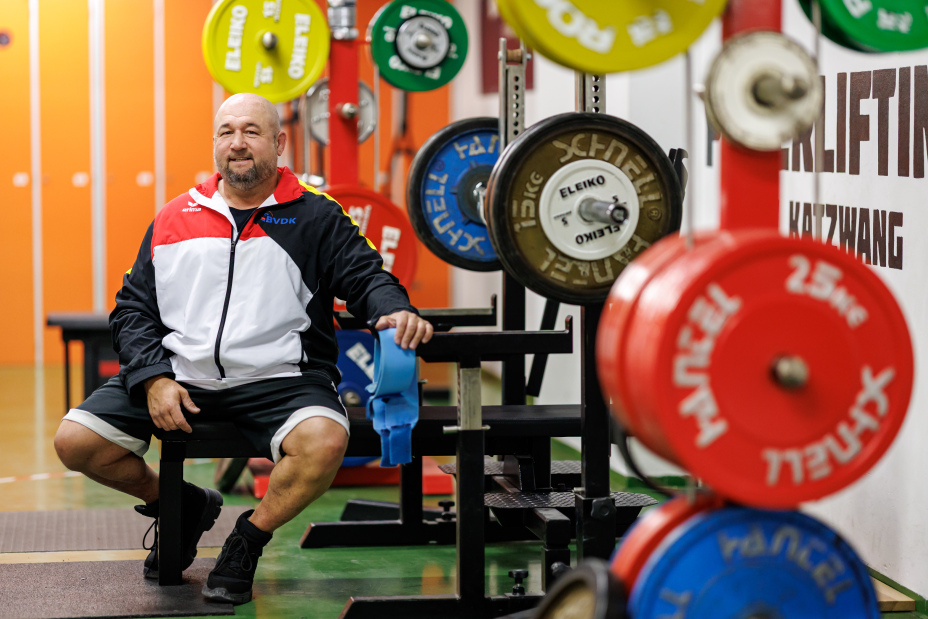 Foto: Kraftsportler Thomas Ziegler sitzt in Trainingsjacke auf einer Bank im Kraftraum. Er ist von bunten Gewichtsscheiben umgeben.