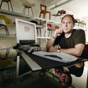 Der Galerist sitzt an seinem Schreibtisch mit PC. Im Hintergrund Stühle junger Designer.