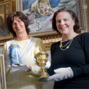 Zwei Frauen zeigen einen Krug und einen Pokal, im Hintergrund hängt ein Gemälde