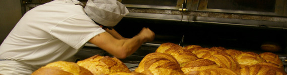 Bäcker schaut in den Ofen, davor liegt ein Blech mit Brötchen.
