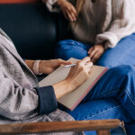 Ein Person schreibt in ein Notizbuch, eine andere Person sitzt auf dem Sofa daneben.